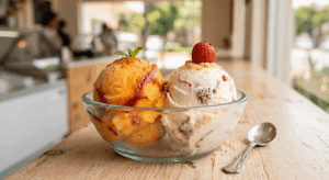 Un primer plano de un tazón de helado con dos sabores: mango y fresa. El helado de mango está decorado con chamoy y Tajín, y el helado de fresa está decorado con trozos de fresa y galletas trituradas. En el fondo, hay una heladería borrosa.